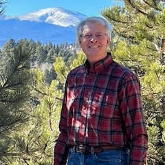 Phil Lockwood smiles at the camera, wearing a flannel button-down shirt and jeans. In the background are pine trees and in the distance a Colorado mountainscape